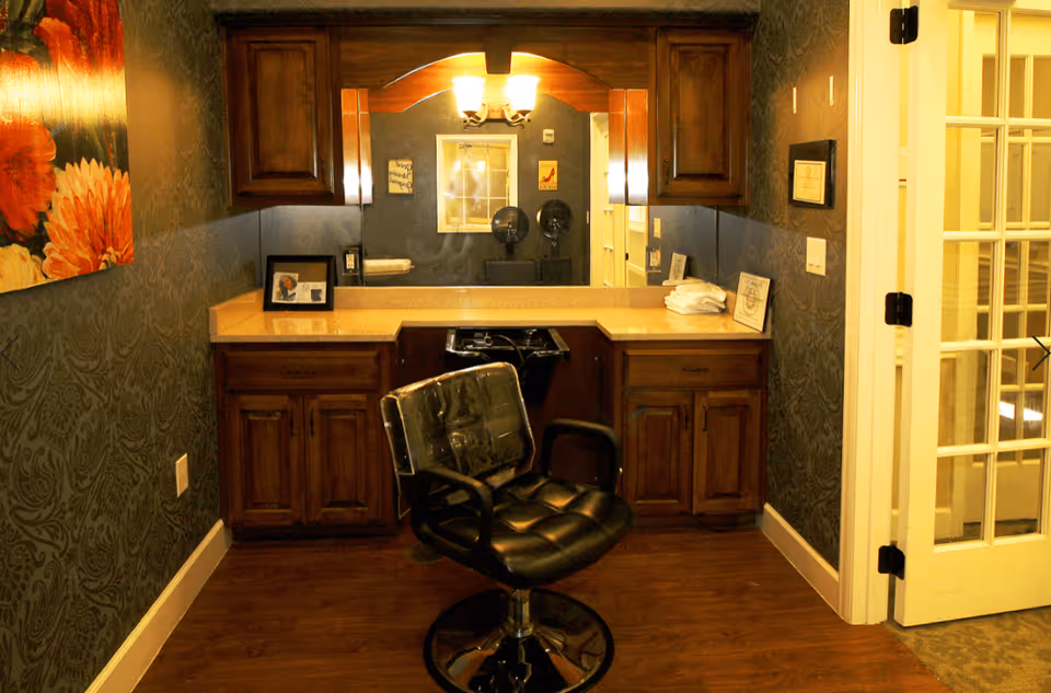 A small salon area in a senior living facility with a black salon chair in front of a countertop with wooden cabinets and a large mirror. The walls have patterned wallpaper, and there is a floral painting on the left wall. A glass-paneled door is open on the right side.