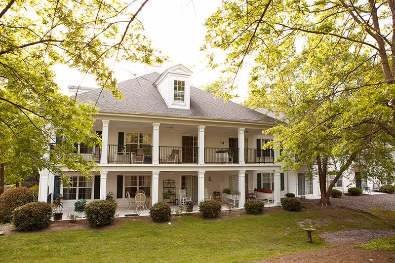 Two-story white building with a covered porch and balcony, surrounded by green trees and bushes, with outdoor seating on both levels.