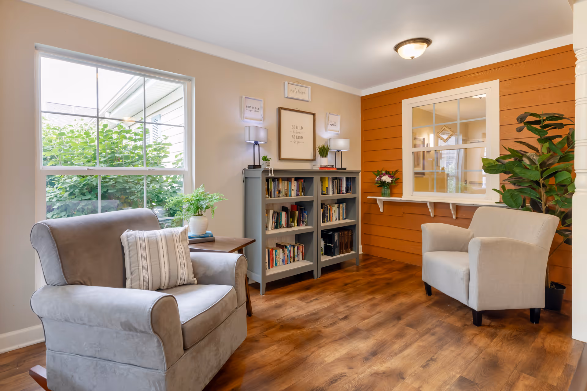 A cozy sitting area with two gray armchairs, a wooden side table with a potted plant, a gray bookshelf filled with books, two table lamps, and decorative framed quotes on the wall. There is a large window showing greenery outside and an interior window with a flower vase on the ledge. The room has warm wooden flooring and an orange accent wall.