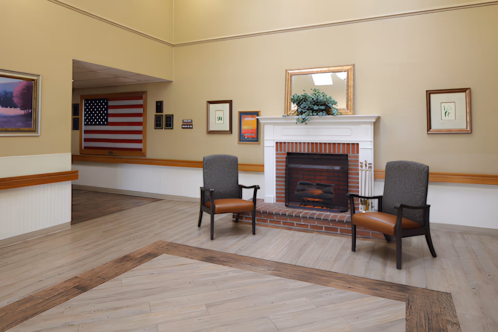 A cozy indoor sitting area with two cushioned chairs facing a white brick fireplace with a decorative plant and a mirror above it. The room has light-colored walls adorned with framed artwork and an American flag visible through a window-like opening. The floor is light wood with a darker wood inlay pattern.