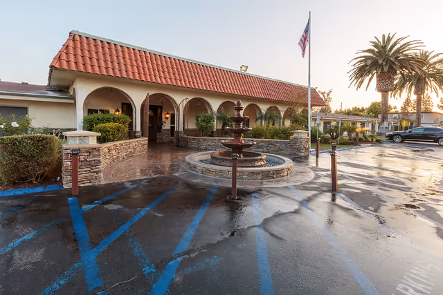 Exterior view of Villa Las Palmas Healthcare Center featuring a building with a red tiled roof, arched entryways, a central water fountain, palm trees, and a parking area with blue handicap markings.
