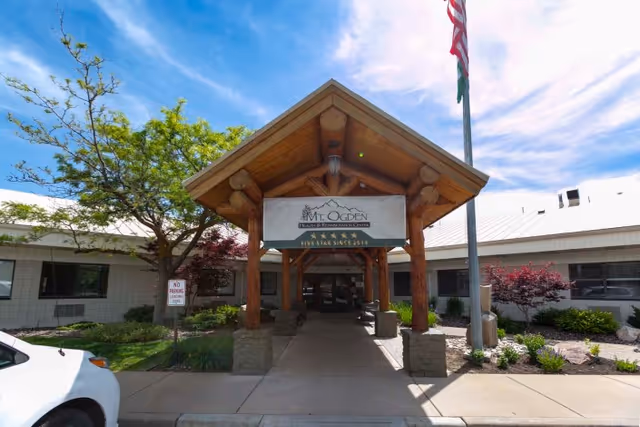 Entrance to Mt. Ogden Health and Rehabilitation Center featuring a wooden covered walkway with stone pillars, an American flag on a flagpole, surrounding greenery, and a clear blue sky with some clouds.