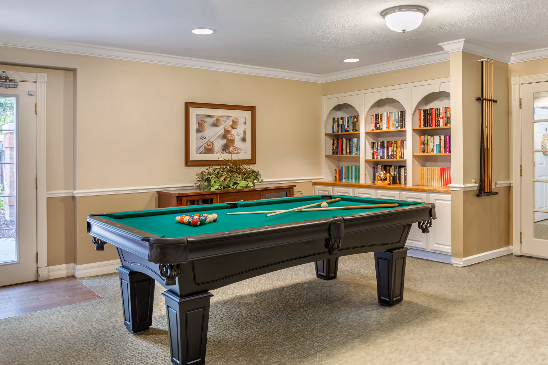 A well-lit room with a pool table in the center, complete with pool cues and balls arranged for a game. Behind the pool table is a wooden cabinet with a plant on top and a framed picture of bingo cards on the wall. To the right, there is a built-in white bookshelf filled with books and a rack holding pool cues. The room has beige walls, carpeted floor, and two glass doors leading outside.