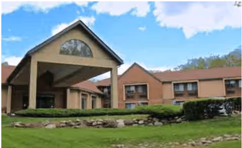 Exterior view of a senior living facility building with a covered entrance, brick walls, multiple windows, and a well-maintained lawn with shrubs and rocks in front under a partly cloudy sky.