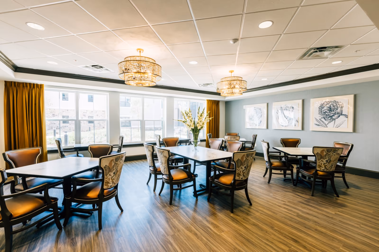 Well-lit communal dining room with multiple tables and upholstered chairs, large windows, chandeliers, and floral wall art.