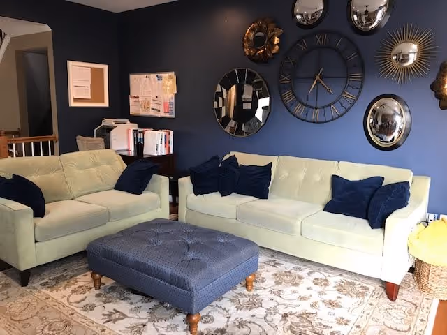 A living room with two pale green sofas, navy blue pillows, a tufted blue ottoman, and decorative mirrors and a large clock on a dark blue accent wall.