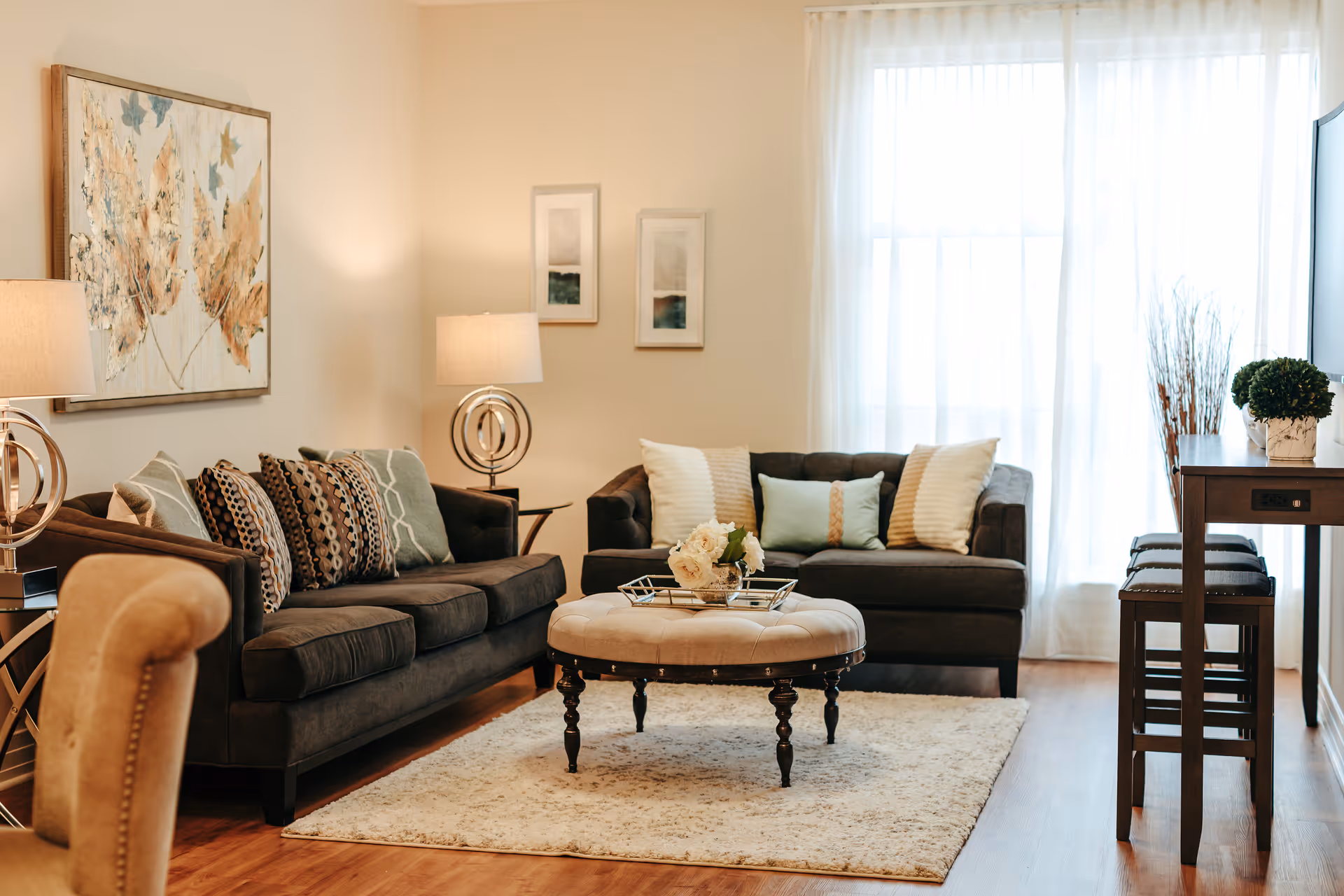 A cozy living room with two dark sofas adorned with multiple decorative pillows, a round tufted ottoman serving as a coffee table with a flower arrangement on top, a light-colored area rug, two modern table lamps with circular bases, framed artwork on the wall, sheer white curtains allowing natural light through a large window, and a tall wooden table with stools on the right side.