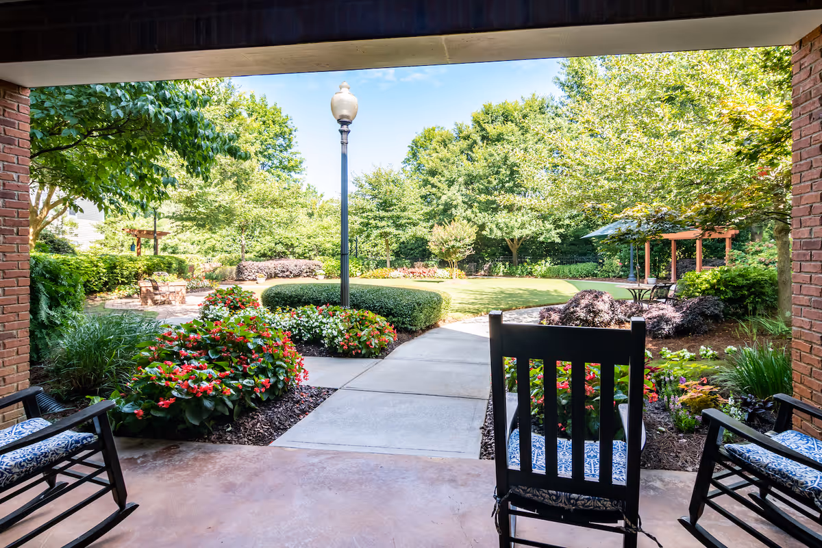View from a covered patio area with two cushioned rocking chairs looking out onto a landscaped garden with a concrete pathway, colorful flower beds, green bushes, trees, a lamppost, and outdoor seating with umbrellas in the background under a clear blue sky.