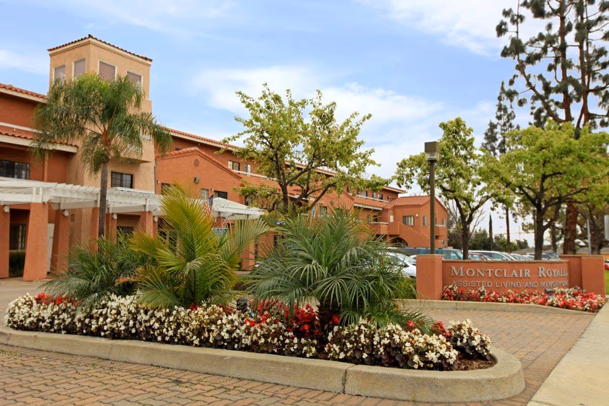 Exterior view of Montclair Royale assisted living and memory care facility showing a landscaped area with palm trees, flowering plants, and a sign with the facility's name. The building is a two-story structure with a terracotta-colored facade and a clear sky above.