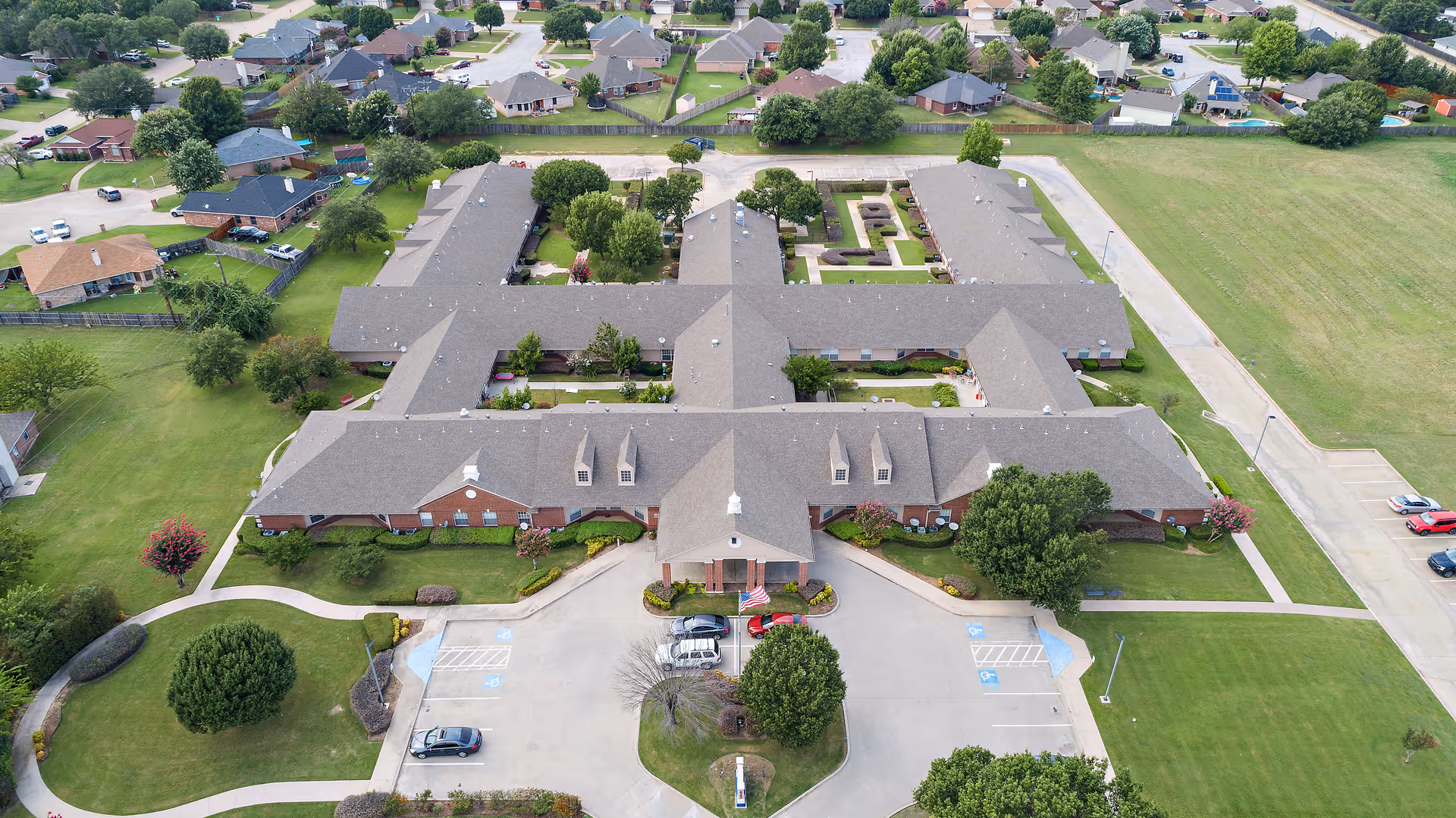 Aerial view of Morada Burleson senior living facility showing a large, single-story building with multiple wings, surrounded by green lawns, trees, and a parking area with several cars. The building has a gray roof and red brick exterior, with pathways and landscaped gardens around it.