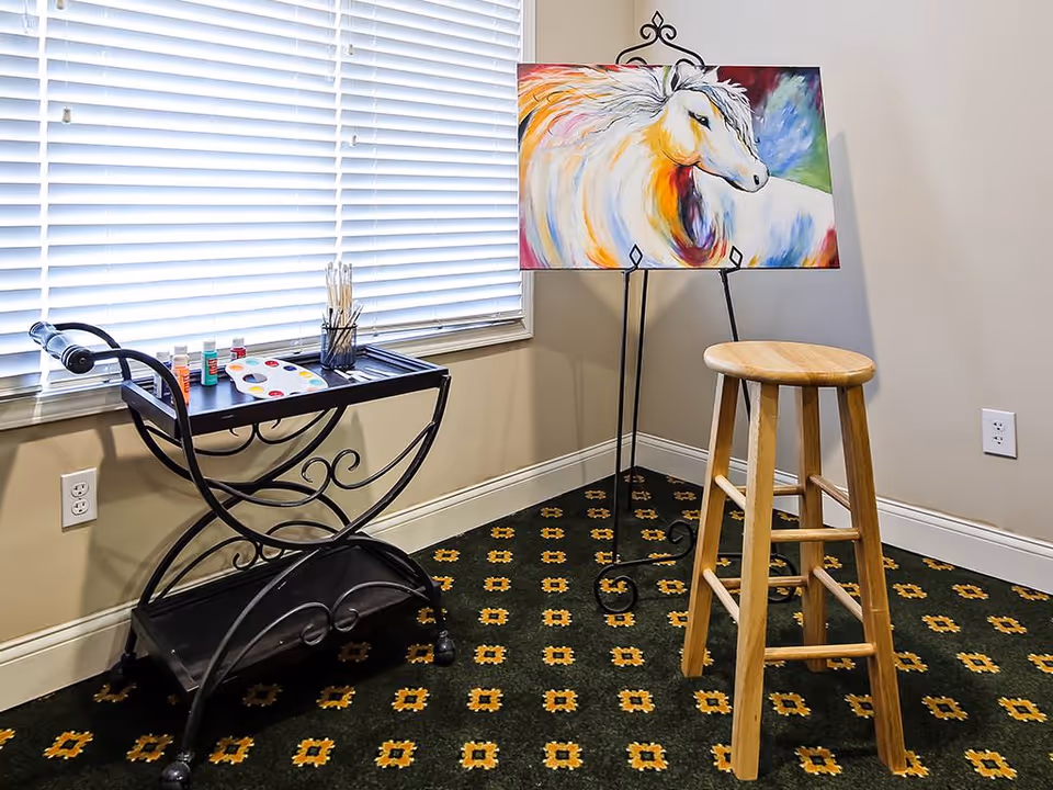 A small art corner in a room with a wooden stool, a colorful painting of a horse on an easel, and a black metal cart holding paint bottles, brushes, and a paint palette. The room has a window with white blinds and patterned carpet flooring.