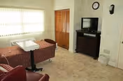 A small room with a hospital bed, a brown recliner chair, a wooden dresser with a television on top, a wall clock, and a window with vertical blinds letting in natural light.