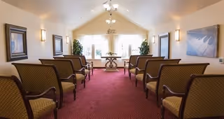 A small chapel or meeting room with rows of cushioned chairs arranged facing a table with a floral arrangement near large windows. The room has a red carpet, cream-colored walls, framed artwork, and wall sconces providing lighting.