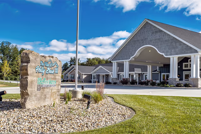 Front entrance of the Villas of Holly Brook assisted living facility with a stone sign, landscaped rock bed, and a covered porte-cochere.