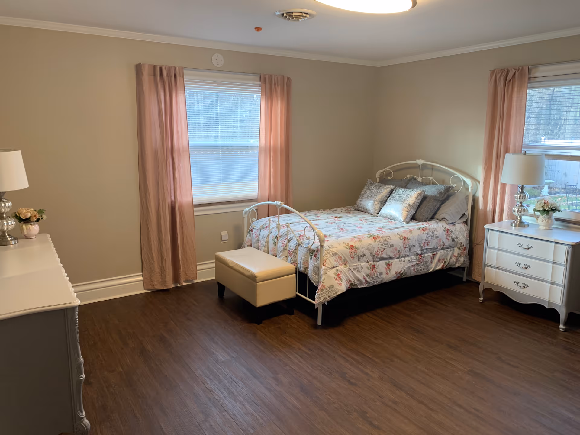 A cozy bedroom with a white metal bed frame, floral bedspread, and several pillows. The room has two windows with pink curtains, a white dresser with a lamp and flower vase on the left, and a white nightstand with a lamp and flower vase on the right. The floor is dark wood, and the walls are painted beige.