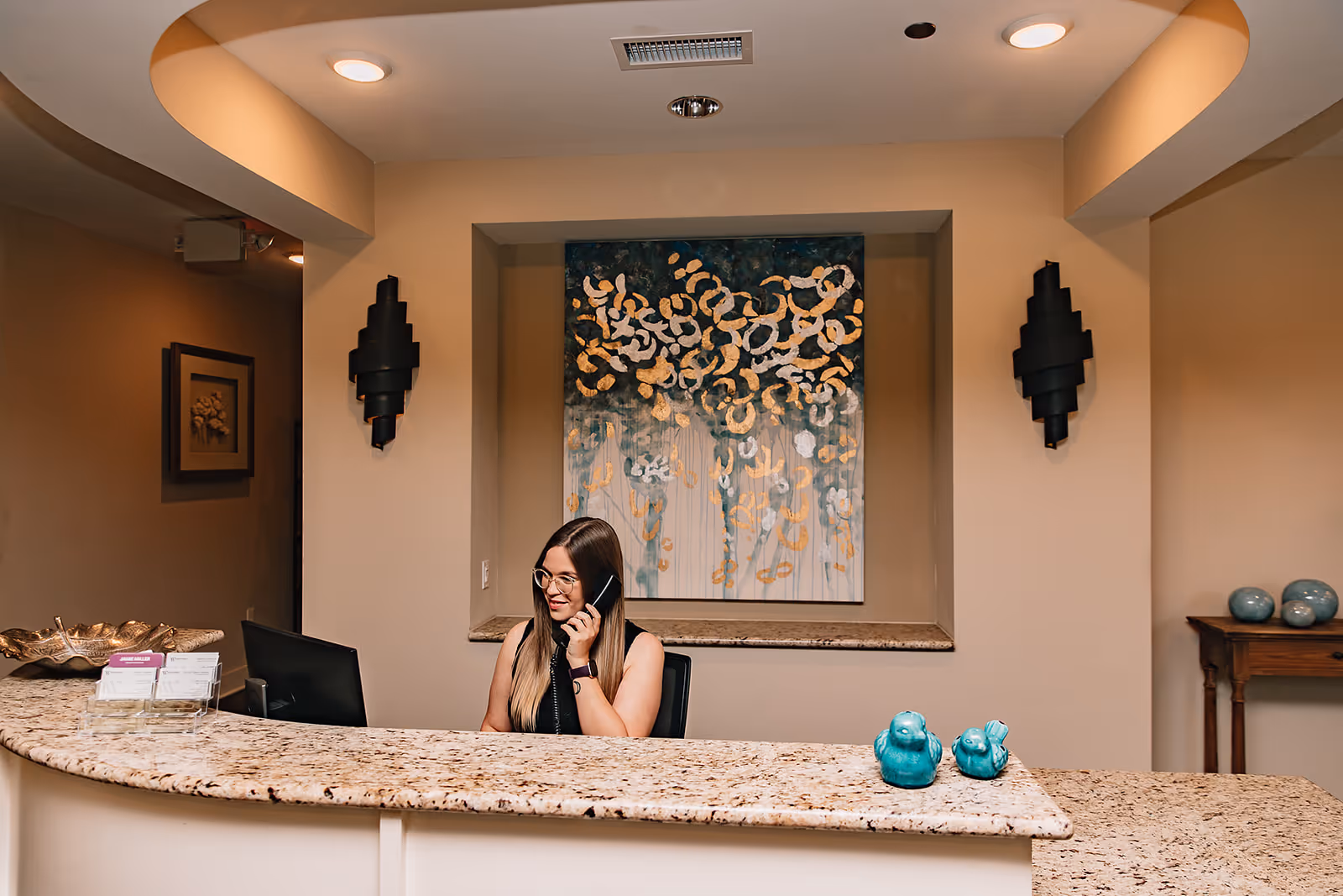 Reception desk inside a senior living facility lobby with a receptionist on the phone, decorative artwork and accents behind the desk.