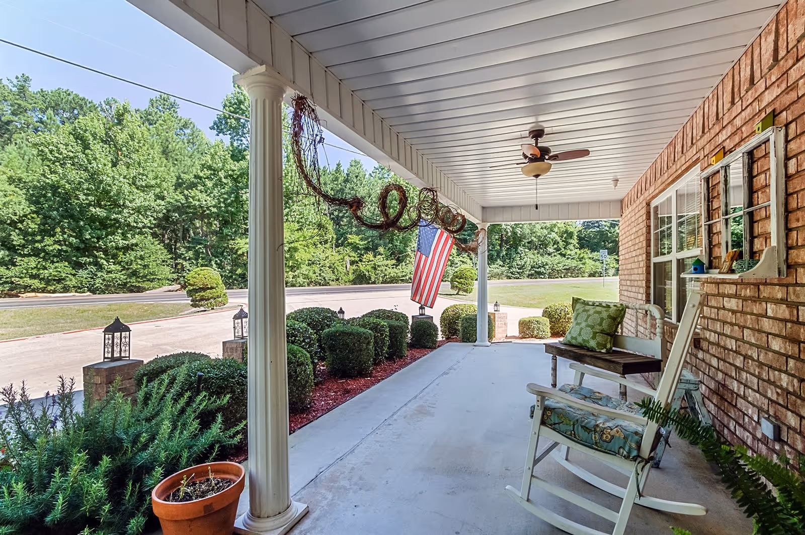 Covered front porch with rocking chairs, columns, an American flag, and trimmed landscaping along a driveway.