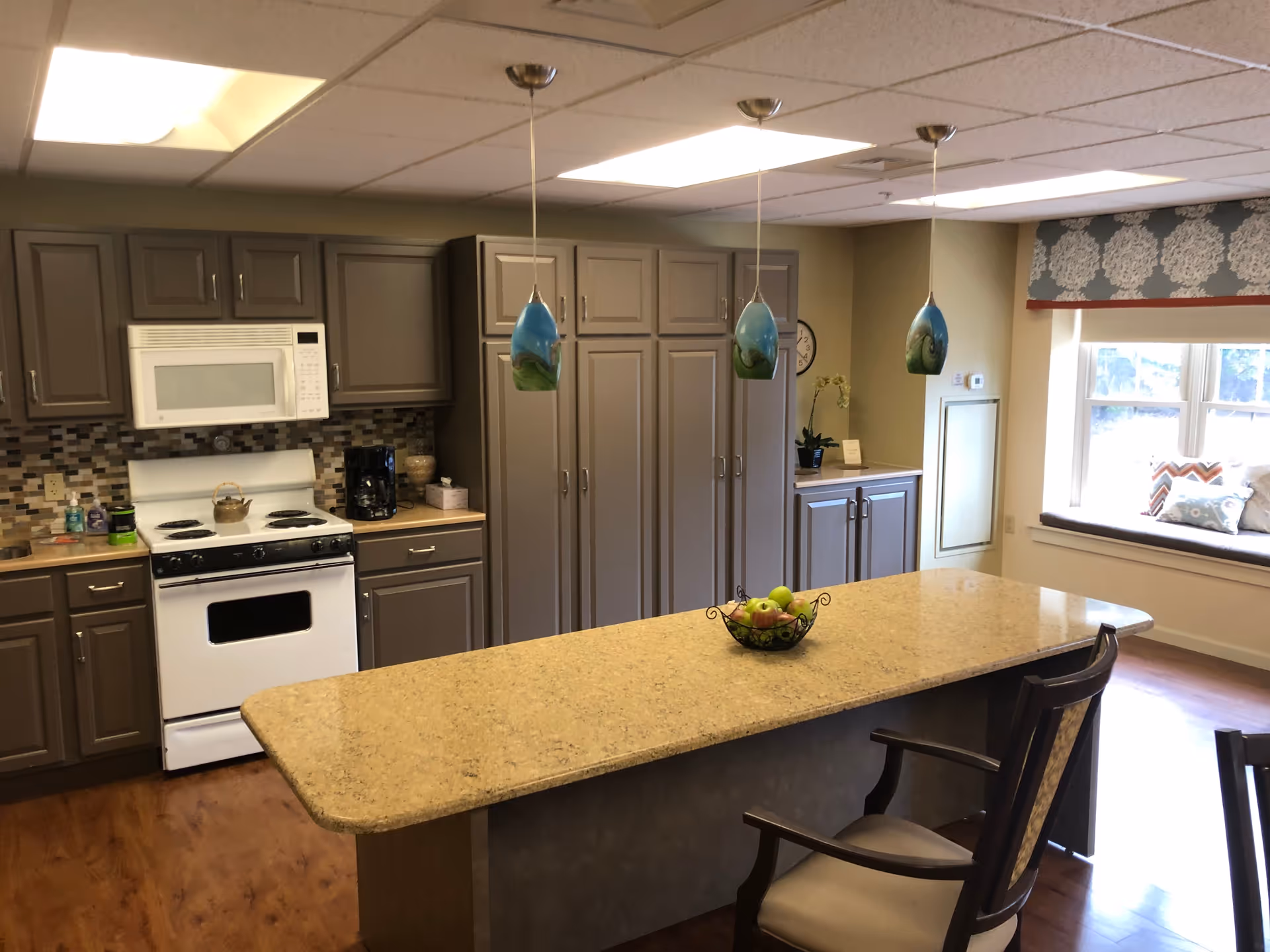 Bright communal kitchen with a large island topped with a fruit bowl, pendant lights, gray cabinets, a stove, and a window seat.