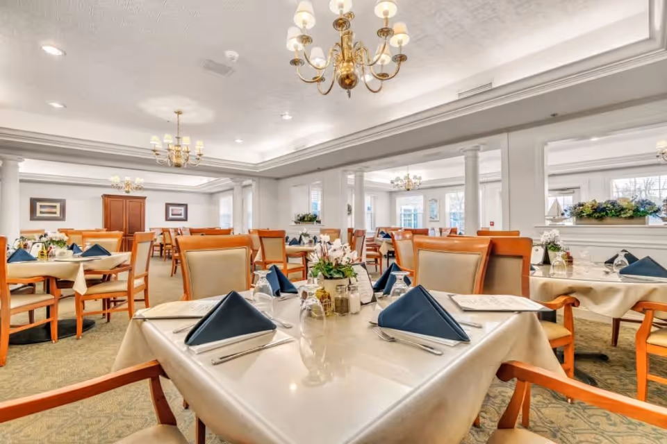 A bright and elegant dining room with multiple tables set with white tablecloths, blue folded napkins, silverware, and glassware. The room features wooden chairs with beige cushions, chandeliers hanging from the ceiling, white columns, and large windows allowing natural light to fill the space. Decorative plants and framed artwork adorn the walls.