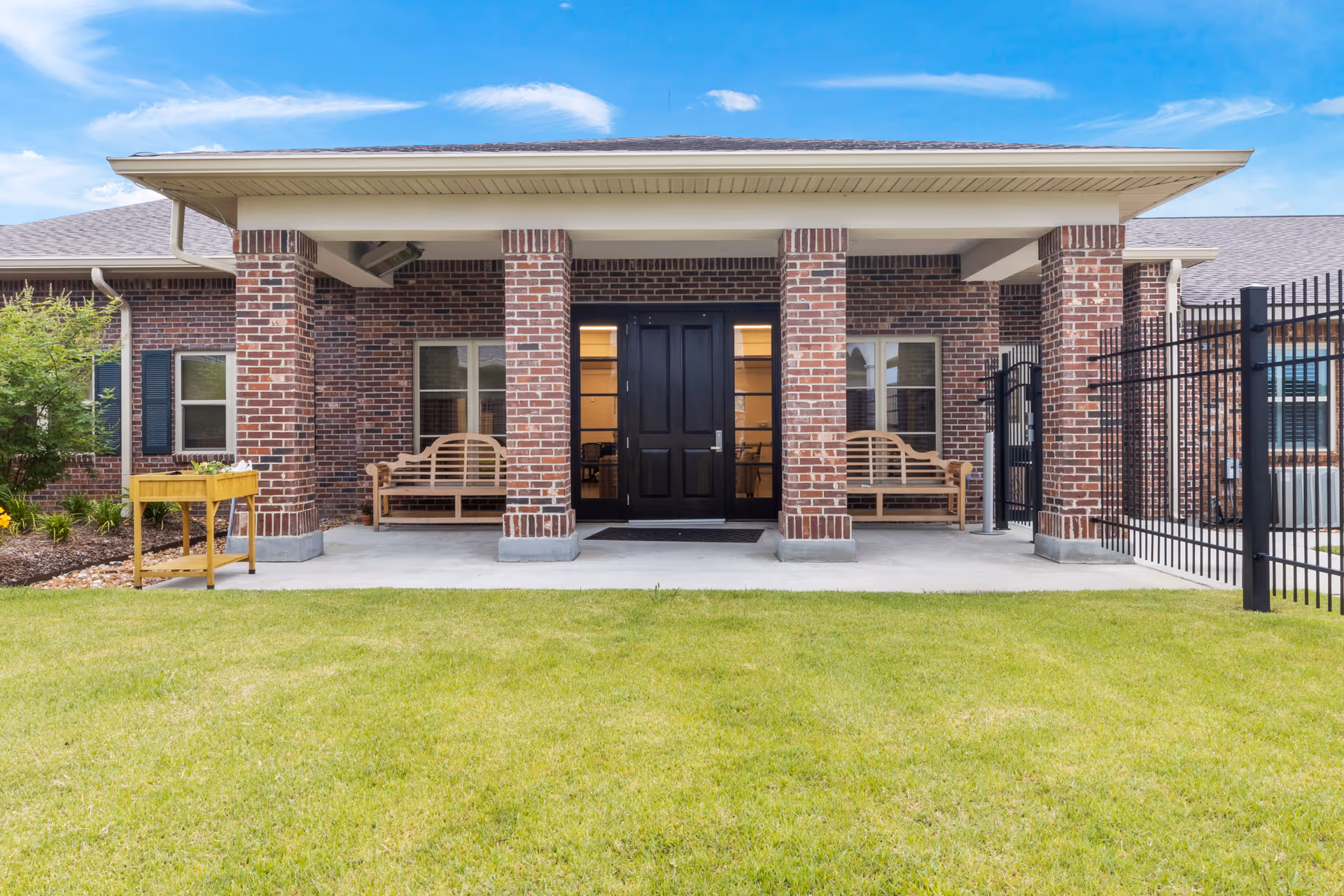 Front view of a brick building with a covered porch supported by four brick columns. The porch has two wooden benches on either side of a black double door entrance. There is a green lawn in the foreground and a clear blue sky with some clouds above.
