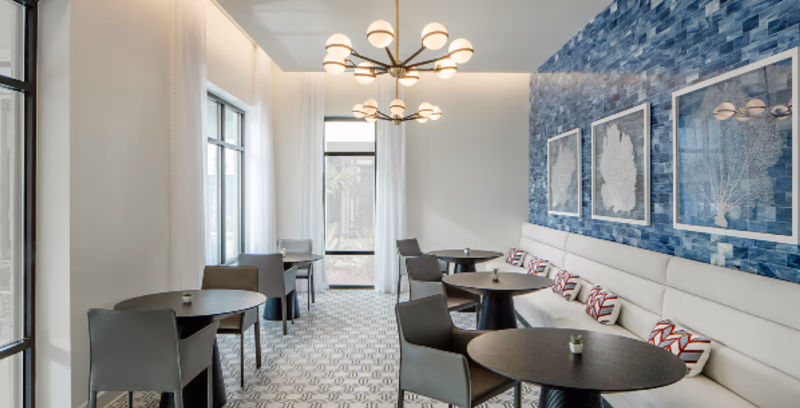 Bright modern dining room with round black tables, gray chairs and banquette seating along a blue-tiled accent wall, lit by pendant chandeliers and large windows.