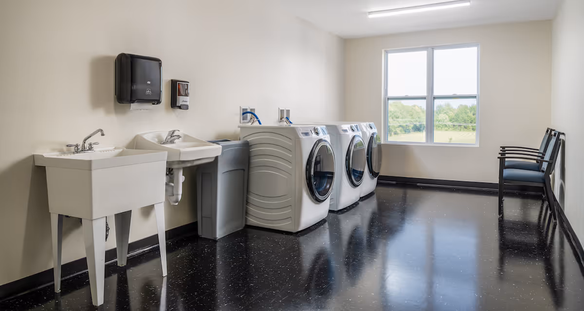 Laundry room with three front-loading washing machines, a utility sink, a handwashing sink, a paper towel dispenser, a soap dispenser, and three chairs lined up against the wall near a window showing greenery outside.