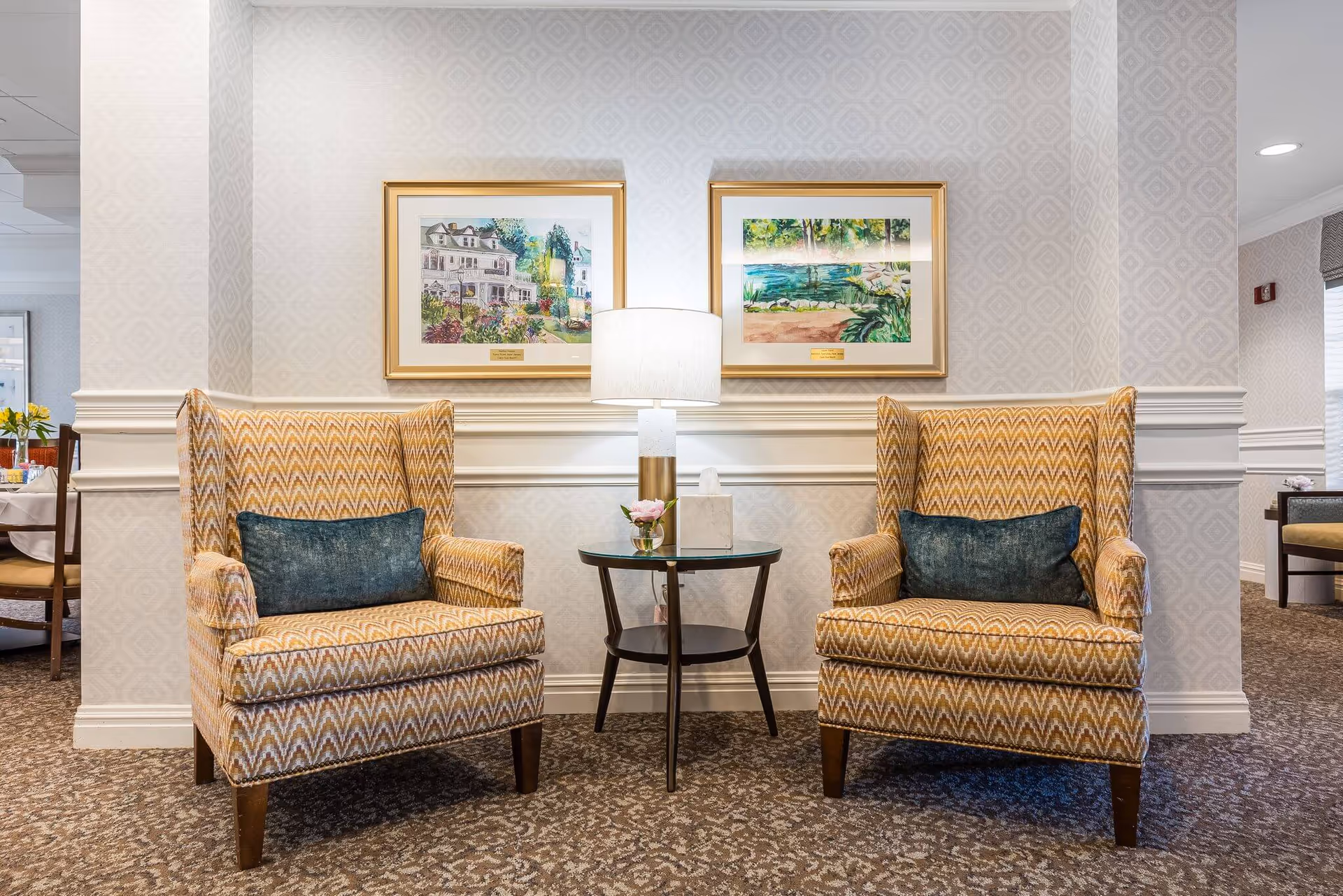 Two patterned armchairs with a small table and lamp between them in a well-lit common area with framed artwork on the wall.