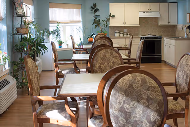 A bright dining area with several wooden tables and chairs featuring patterned upholstery. The room has large windows letting in natural light, potted plants on the windowsill and floor, and a kitchen area with white cabinets and a black stove in the background.