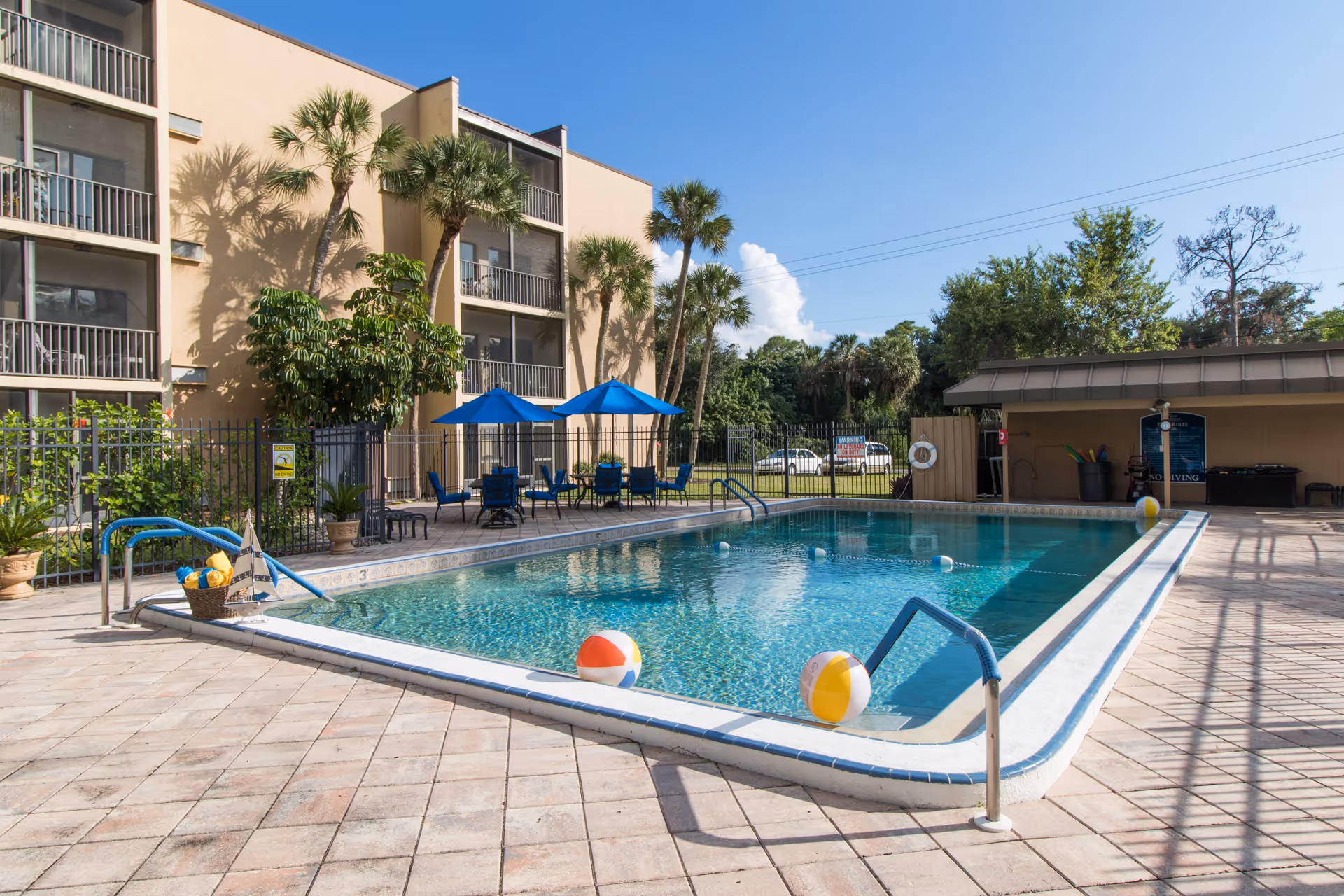 Outdoor swimming pool area at River Commons with clear blue water, colorful beach balls floating in the pool, blue patio umbrellas, and chairs on the pool deck. The pool is surrounded by a fence and palm trees, with a beige multi-story building in the background under a clear blue sky.