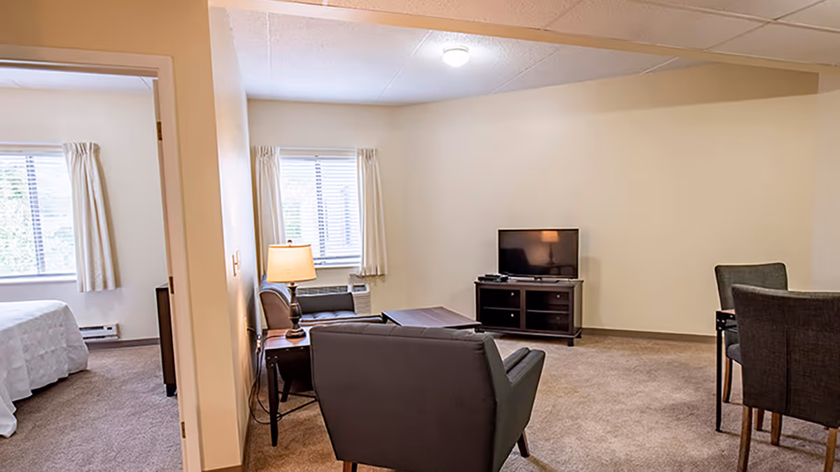 A living room area with beige walls and carpet. The room contains a TV on a stand, a coffee table, two armchairs, a side table with a lamp, and a small dining table with chairs. A bedroom with a bed and window with curtains is visible through an open doorway.