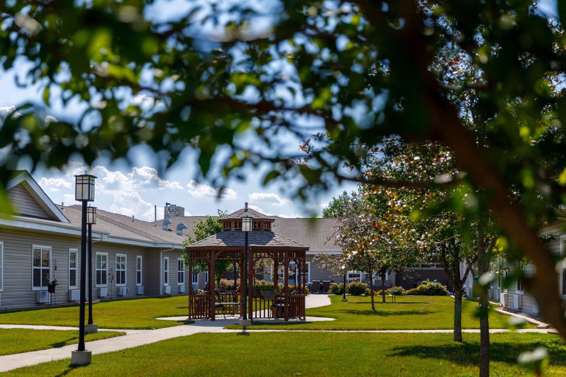 Outdoor courtyard area of a senior living facility with a wooden gazebo at the center, surrounded by green grass, trees, and paved walkways. The facility buildings with multiple windows are visible in the background under a partly cloudy blue sky.