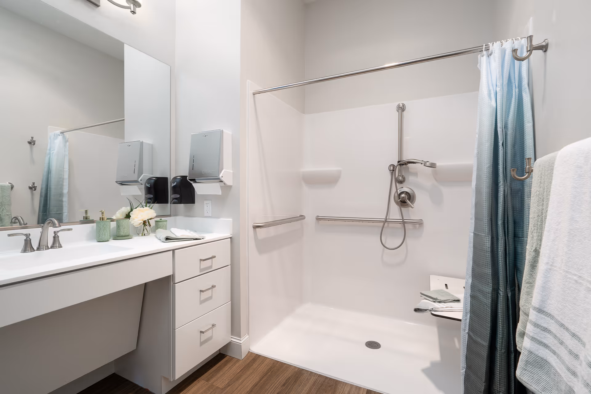 Accessible bathroom with a walk-in shower featuring grab bars and a handheld showerhead, plus a vanity with sink, mirror and towels.