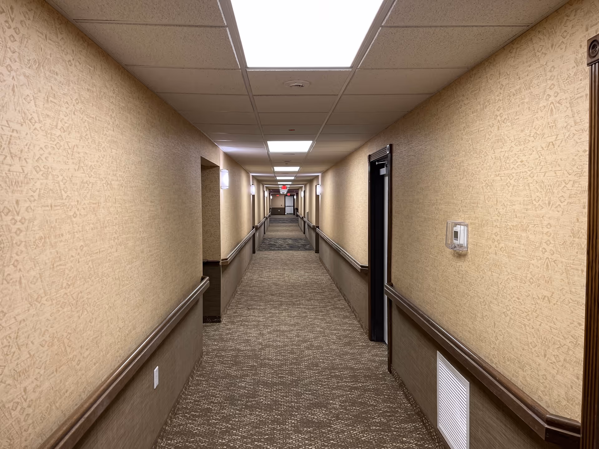 Long indoor hallway with beige patterned walls, carpeted floor, and ceiling lights. Handrails run along both sides of the hallway, with several doorways visible on the right side.