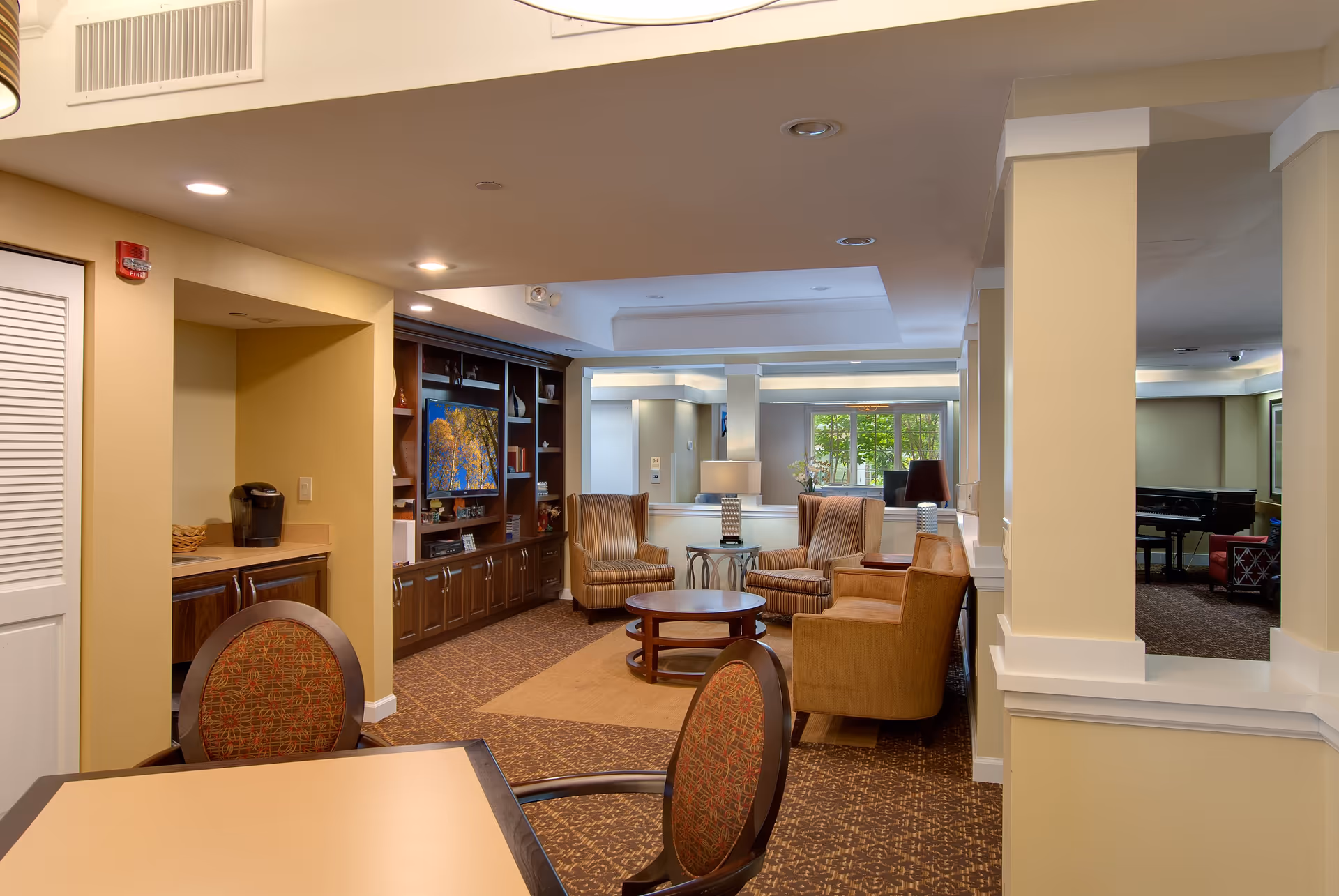 A cozy and well-lit common area in a retirement community featuring a seating arrangement with armchairs and a round coffee table. There is a built-in wooden entertainment center with a television and decorative items. The room has beige walls, carpeted floors, and a window letting in natural light. A small kitchenette area with a coffee maker is visible to the left, and a piano can be seen in the background to the right.