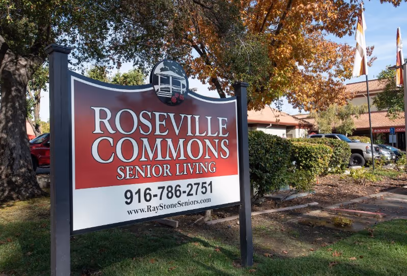 Front exterior view of Roseville Commons Senior Living sign with building, trees, and parked cars.