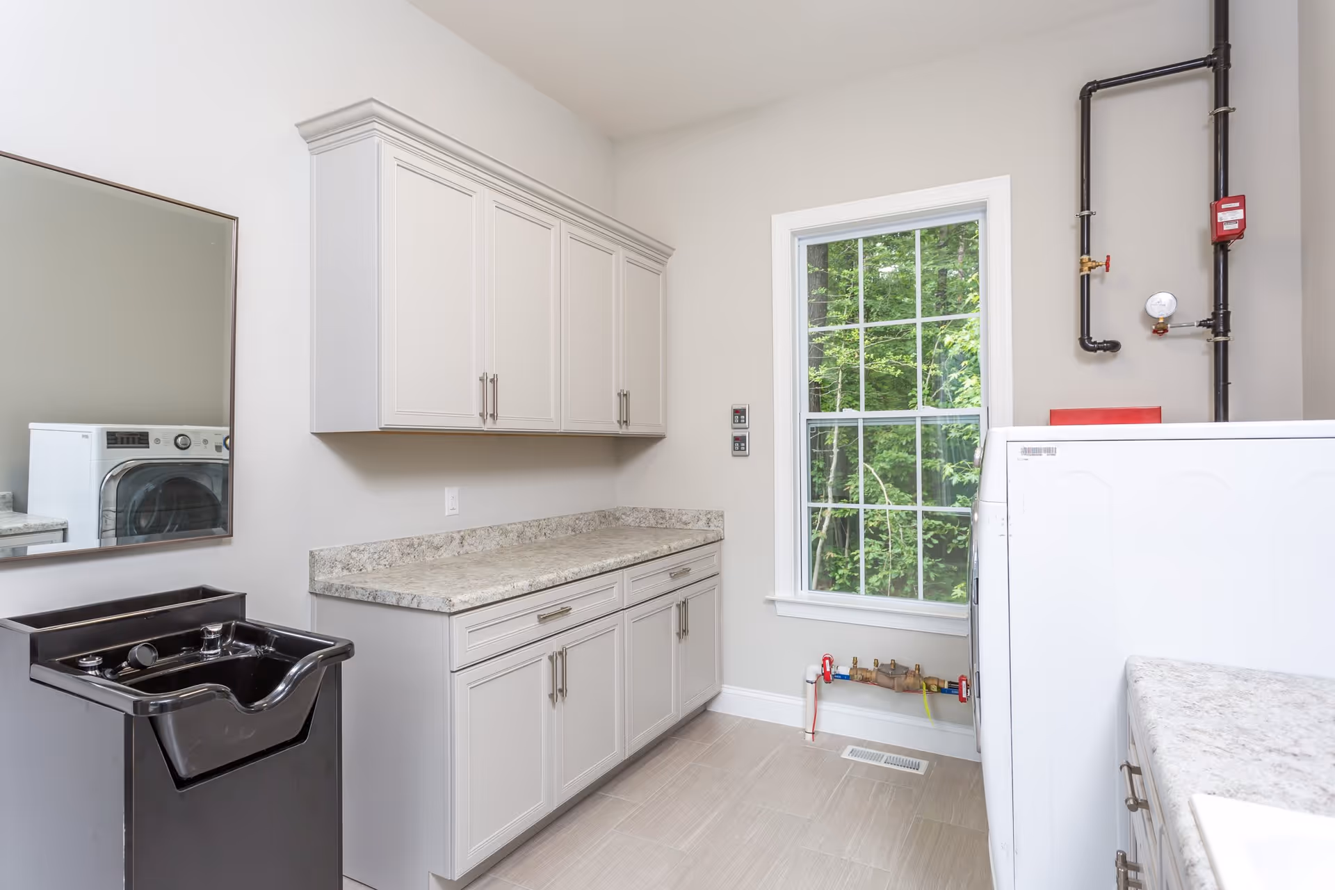 A bright laundry room with white cabinets, a countertop, a black utility sink, a washing machine, and a window showing green trees outside. There are exposed pipes and a pressure gauge on the wall.