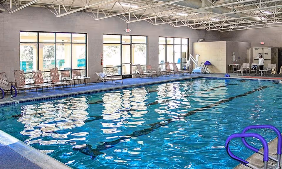 Indoor swimming pool with lane markings, lounge chairs along the side and large windows letting in daylight.
