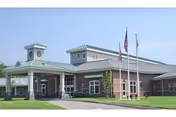 Exterior view of a single-story brick building with a covered entrance, multiple windows, and two flagpoles displaying the American flag and another flag. The building has a light green roof with a small tower-like structure on top.