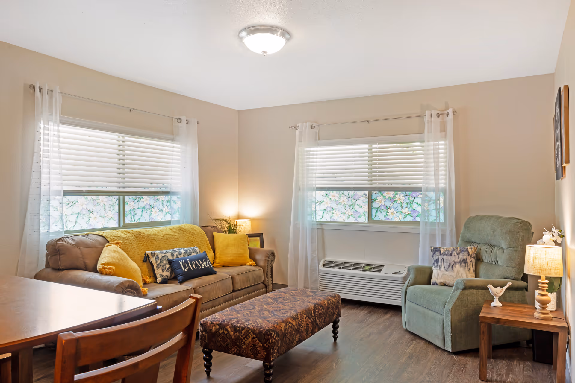 A cozy living room with a brown sofa adorned with yellow and patterned pillows, a green armchair with a decorative pillow, a patterned ottoman, and a wooden side table with a lamp and small decorative items. The room has two windows with white sheer curtains and floral window films, light beige walls, and wood flooring.