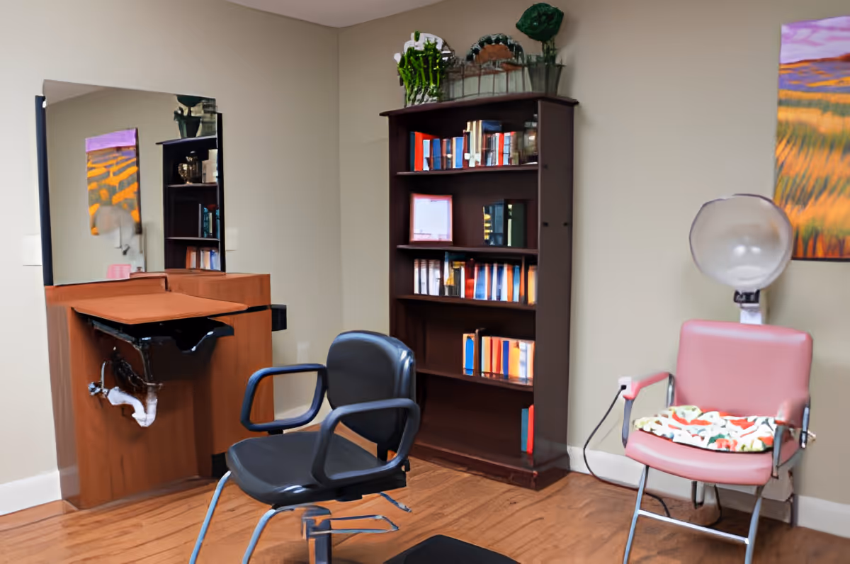 Interior of a salon area in a senior living facility with a black salon chair in front of a wooden sink station with a mirror. To the right, there is a pink salon chair with a hair dryer hood attached, and a colorful cushion on the seat. A tall wooden bookshelf filled with books and decorative items is against the wall. A colorful painting hangs on the wall above the pink chair.