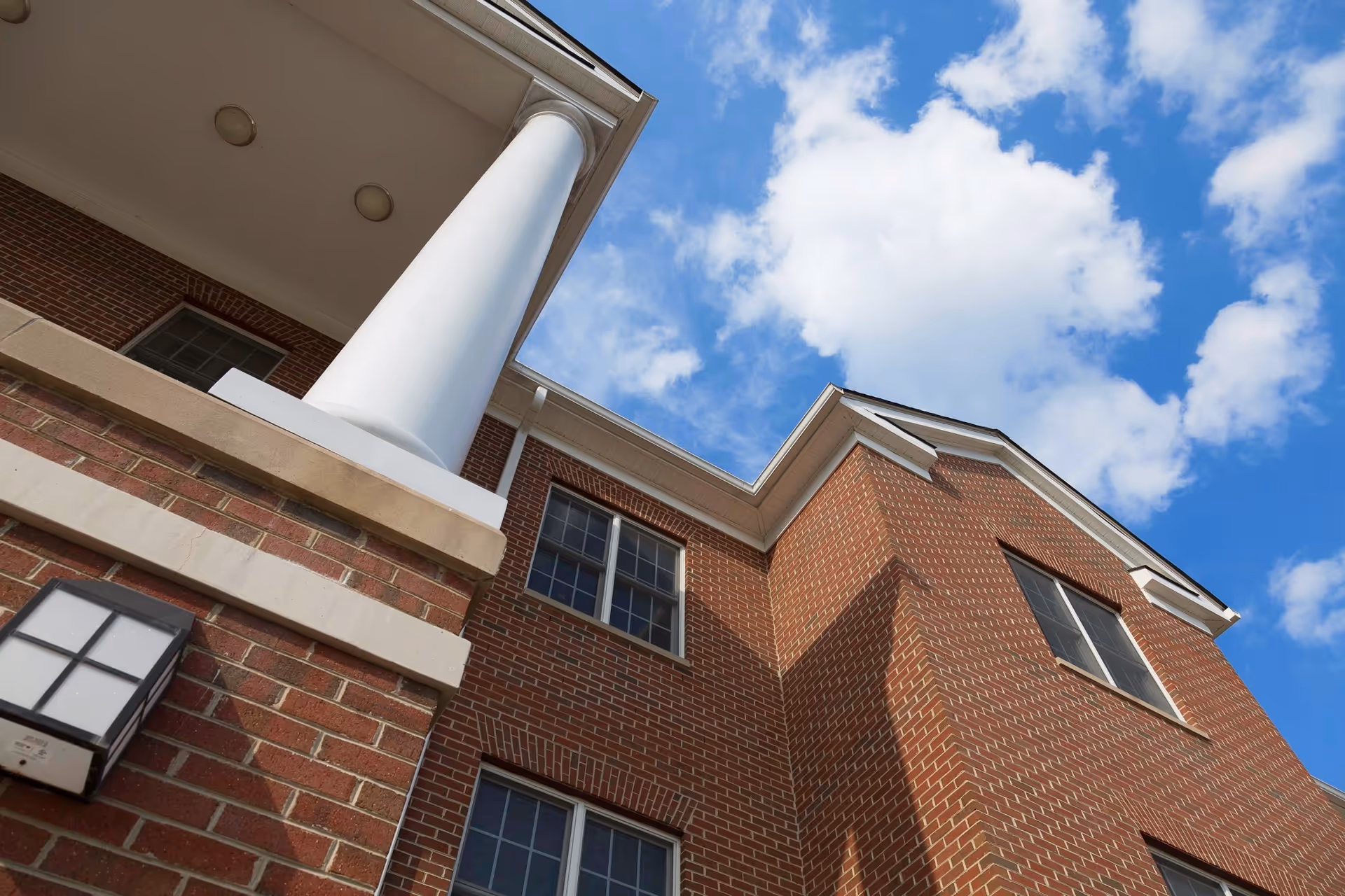 Upward view of a brick building with white columns and multiple windows under a blue sky with scattered clouds.