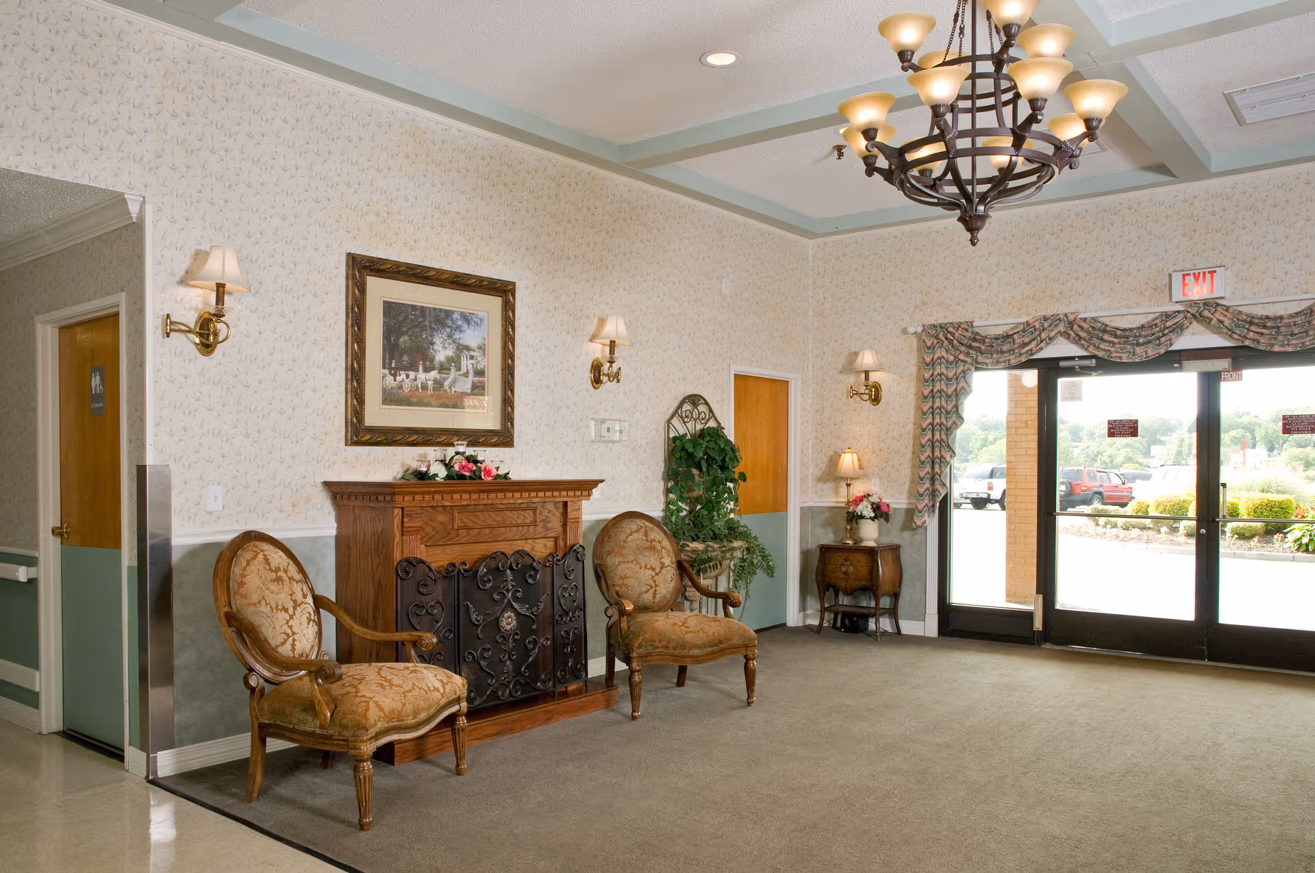 A well-lit interior common area with two upholstered armchairs placed on either side of a decorative wooden fireplace with a black ornate screen. Above the fireplace hangs a framed picture. The walls are light-colored with a subtle pattern, and there are wall sconces with lampshades providing additional lighting. A chandelier hangs from the ceiling. To the right, there is a glass double door exit with an exit sign above it, and outside the doors, cars and greenery are visible. A small wooden side table with a lamp and flowers is positioned near the door.