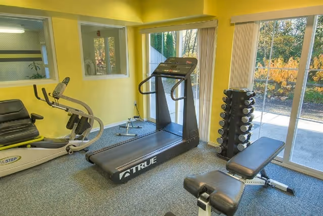 A small fitness room with exercise equipment including a recumbent bike, a treadmill, a weight bench, and a rack of dumbbells. The room has yellow walls, carpeted floor, and large windows with vertical blinds letting in natural light and showing an outdoor view.