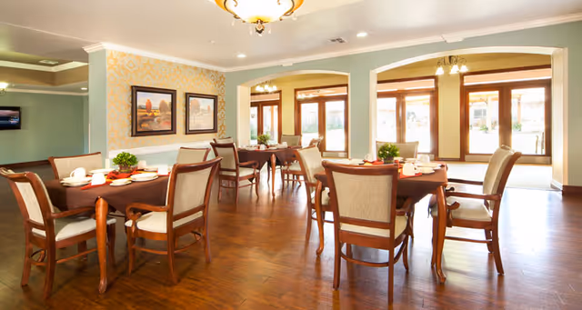 A bright and spacious dining room with wooden floors and several tables covered with brown tablecloths. Each table is set with white plates, cups, and utensils, and has a small green plant centerpiece. The room features large windows and glass doors allowing natural light to fill the space. The walls are decorated with framed artwork and light-colored wallpaper.