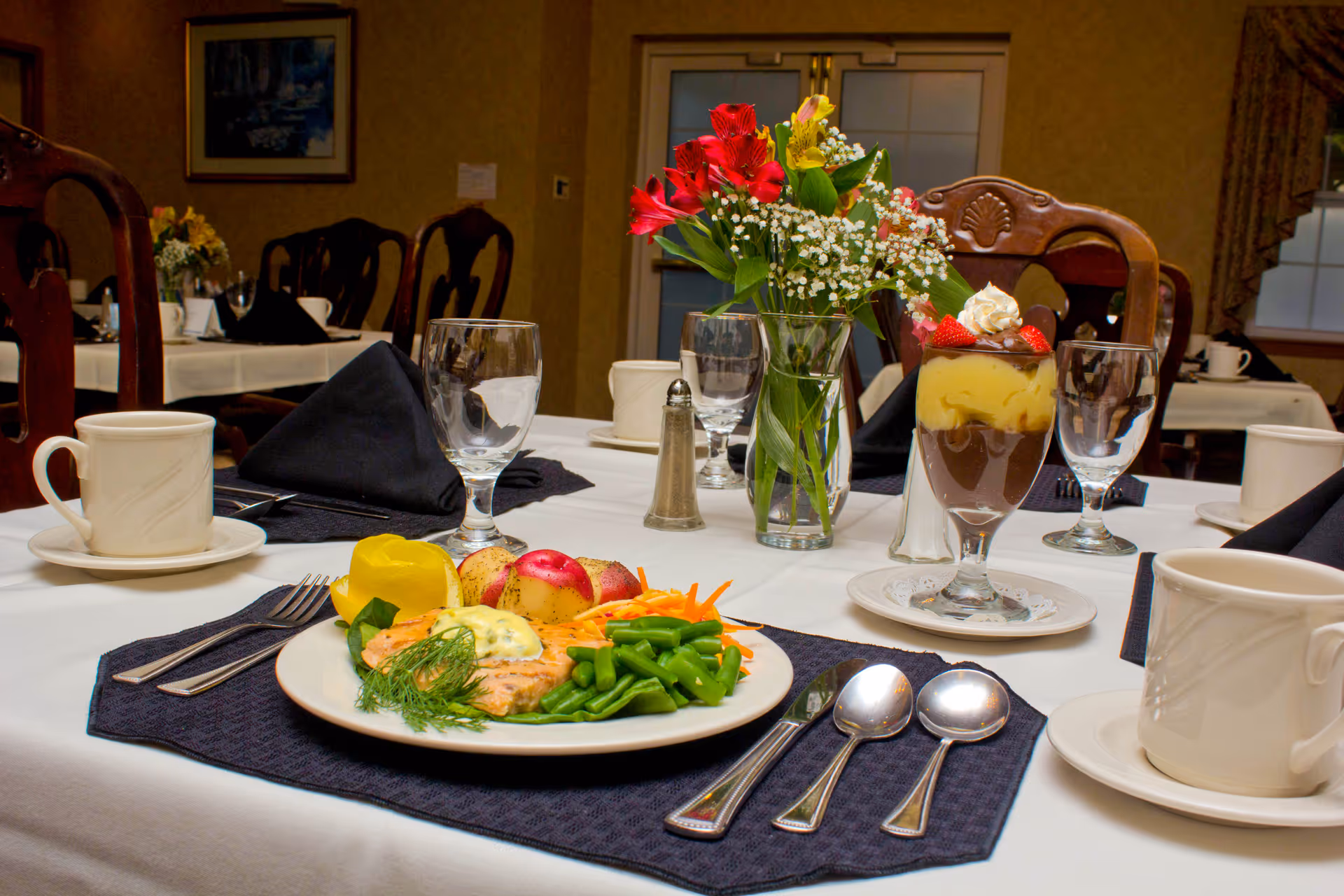 A neatly set dining table with a plated entrée, a dessert parfait, glassware, coffee cups, utensils, and a vase of flowers in a formal dining room.