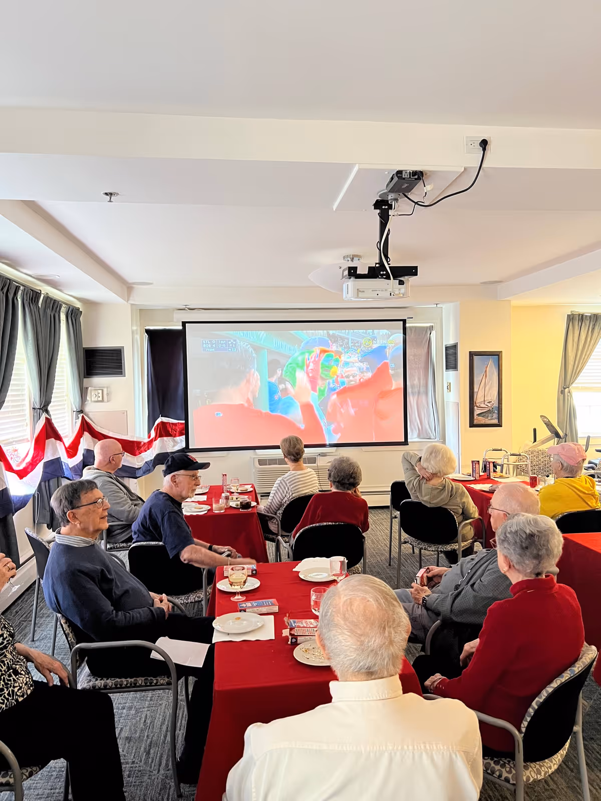 A group of elderly people seated around tables with red tablecloths in a common room, watching a sports event projected on a screen at the front of the room. The room has windows with curtains and patriotic red, white, and blue bunting along the wall.