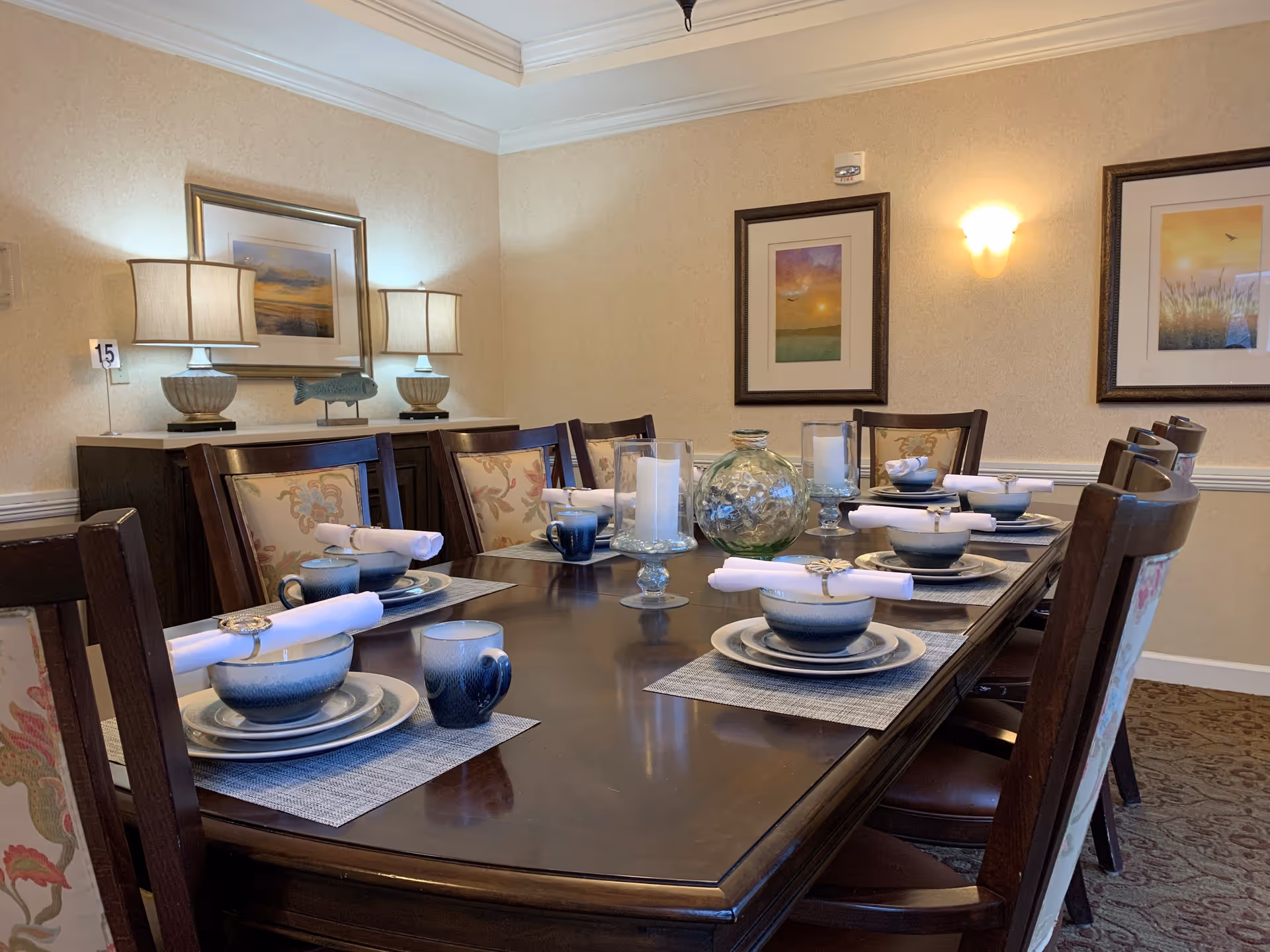 A formal dining room with a long dark wooden table set for eight people. Each place setting includes a blue and white bowl, plate, mug, and a rolled white napkin with a napkin ring. The table also features decorative glass candle holders and a large glass vase centerpiece. The room has beige walls adorned with framed artwork, two table lamps on a sideboard, and a wall sconce providing warm lighting.