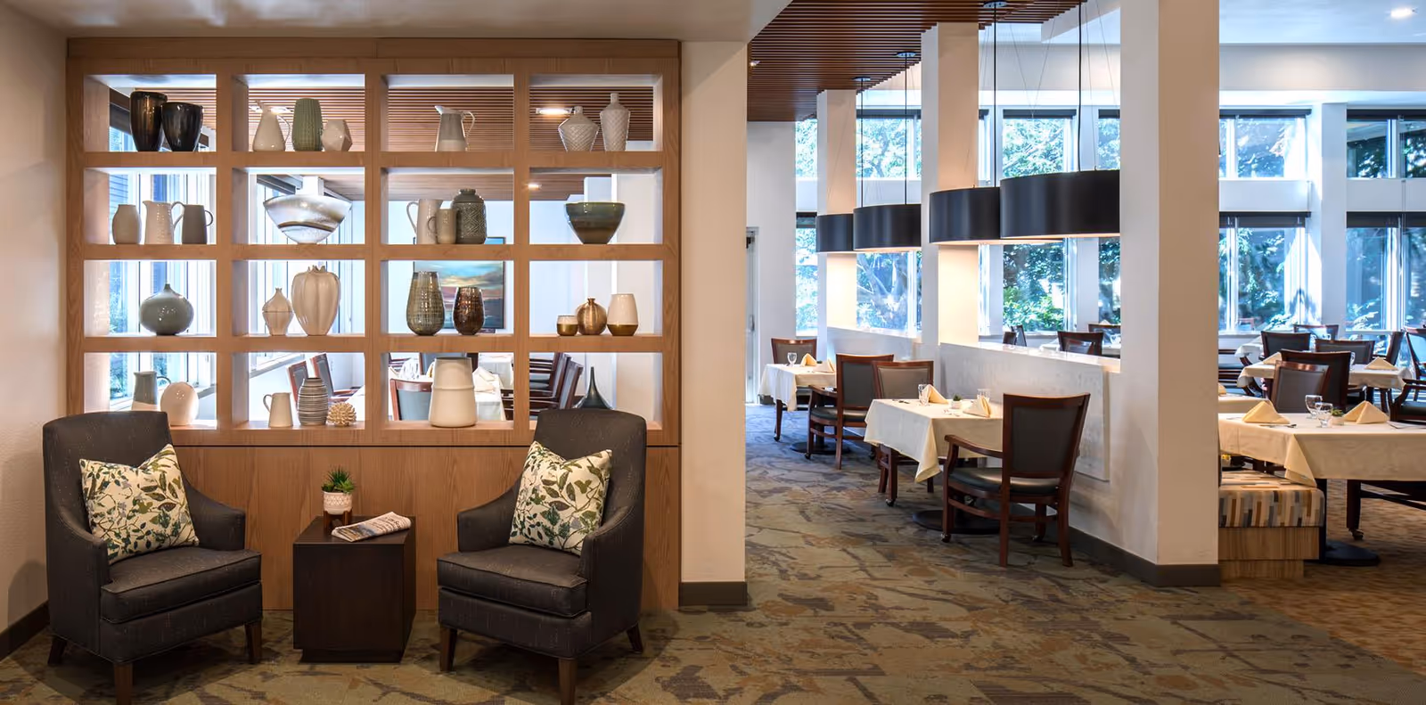 A cozy seating area with two dark armchairs featuring floral cushions and a small wooden table with a plant and a newspaper. Behind the seating area is a wooden partition with decorative vases and pottery. To the right, there is a dining area with tables covered in white tablecloths, wooden chairs, and large black pendant lights hanging from the ceiling. Large windows allow natural light to fill the space.