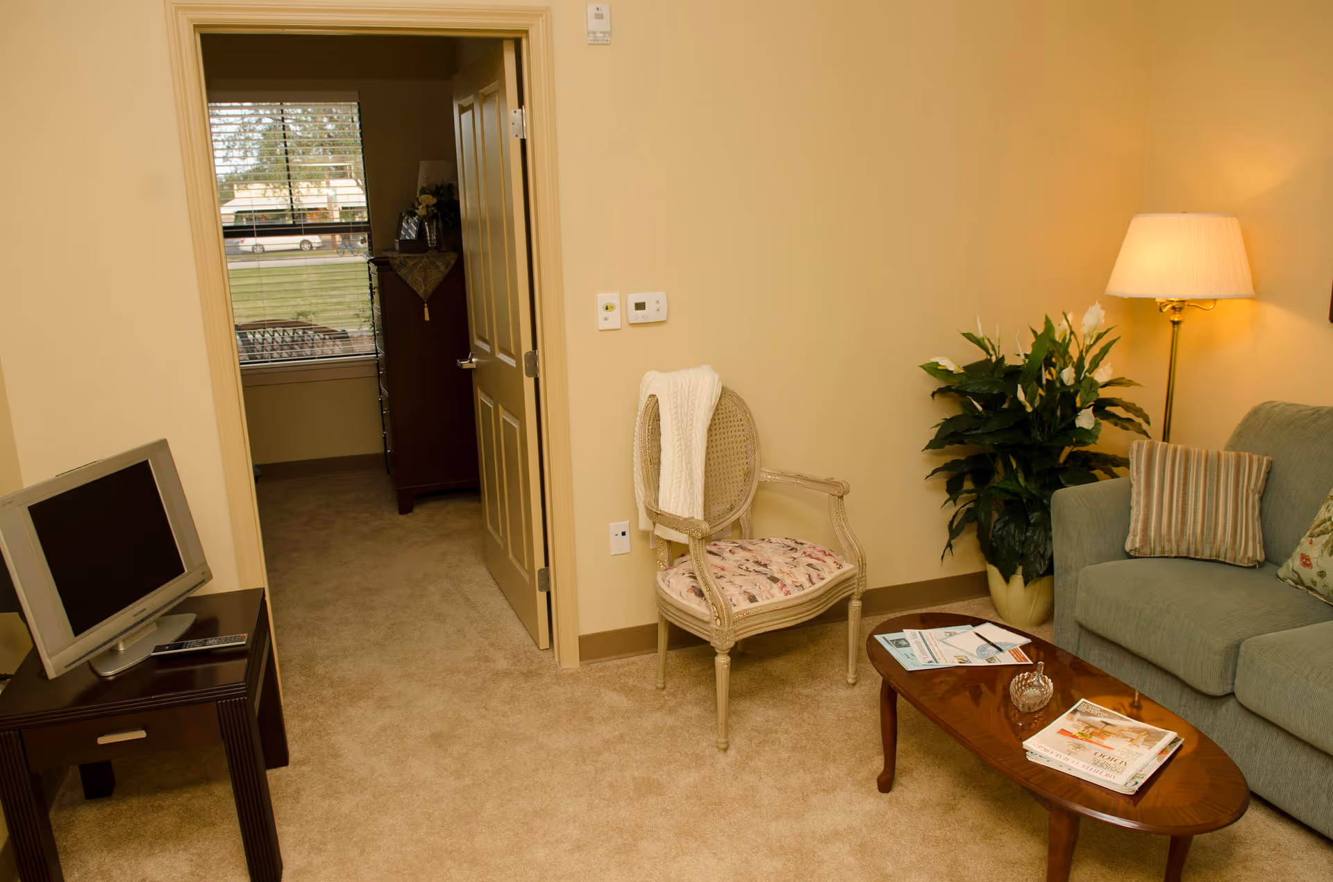 A cozy living room area with beige walls and carpet. There is a green sofa with striped and floral pillows, a wooden coffee table with magazines and a decorative item, a vintage-style chair with a white knitted throw draped over it, a tall floor lamp with a white shade, and a large green potted plant. A doorway leads to another room with a window showing a view of the outdoors and a wooden dresser inside.
