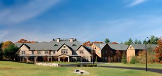 Wide exterior view of a large senior living facility building with multiple peaked roofs, surrounded by green lawns and trees with autumn foliage under a clear blue sky.